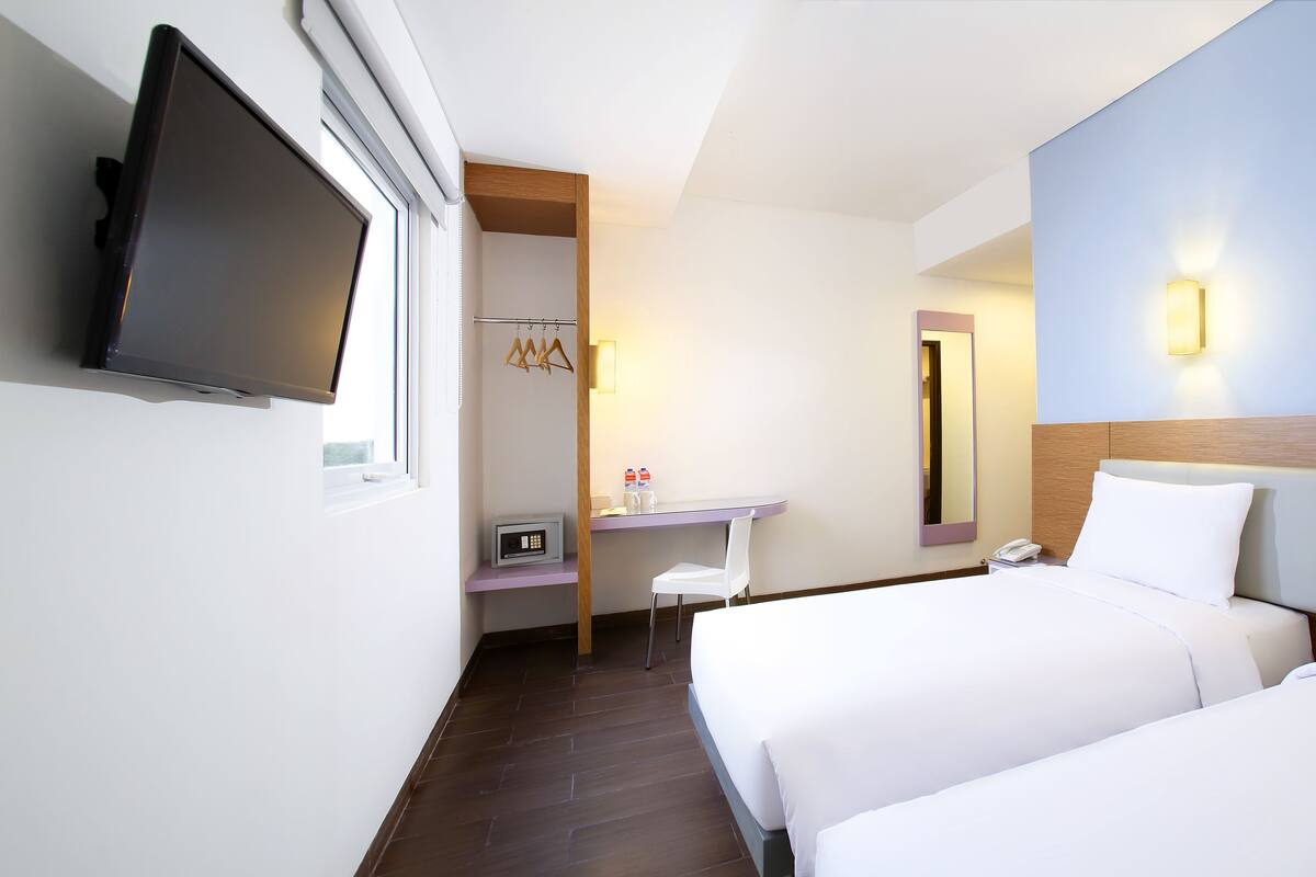 Quarto de hotel com duas camas, televisão fixada na parede, secretária com espelho e um espaço de casa de banho pequeno.
