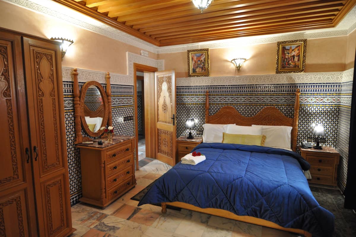 A traditional Moroccan-style bedroom with a wooden bed, a wooden wardrobe, a mirror, and patterned tile walls.