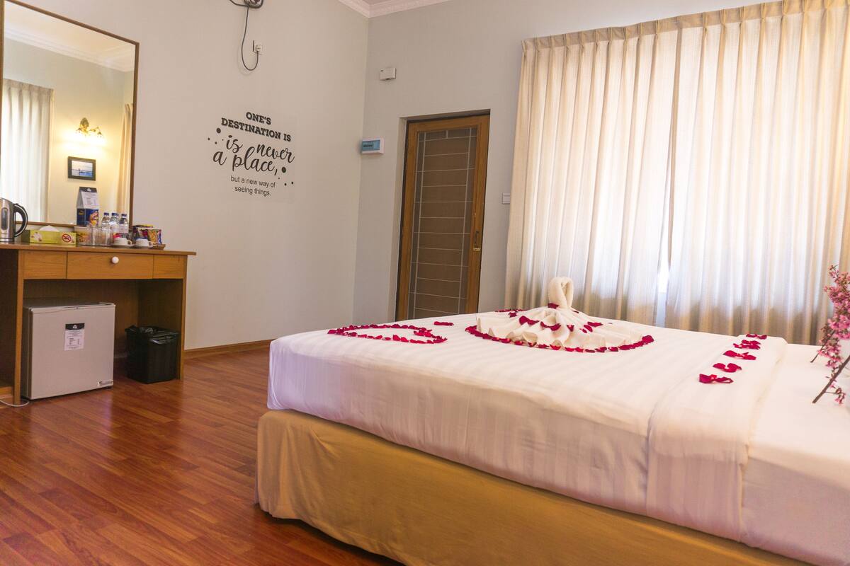 A neatly made bed with rose petals, a wooden desk with a kettle, a small fridge, and a wall with a motivational quote.
