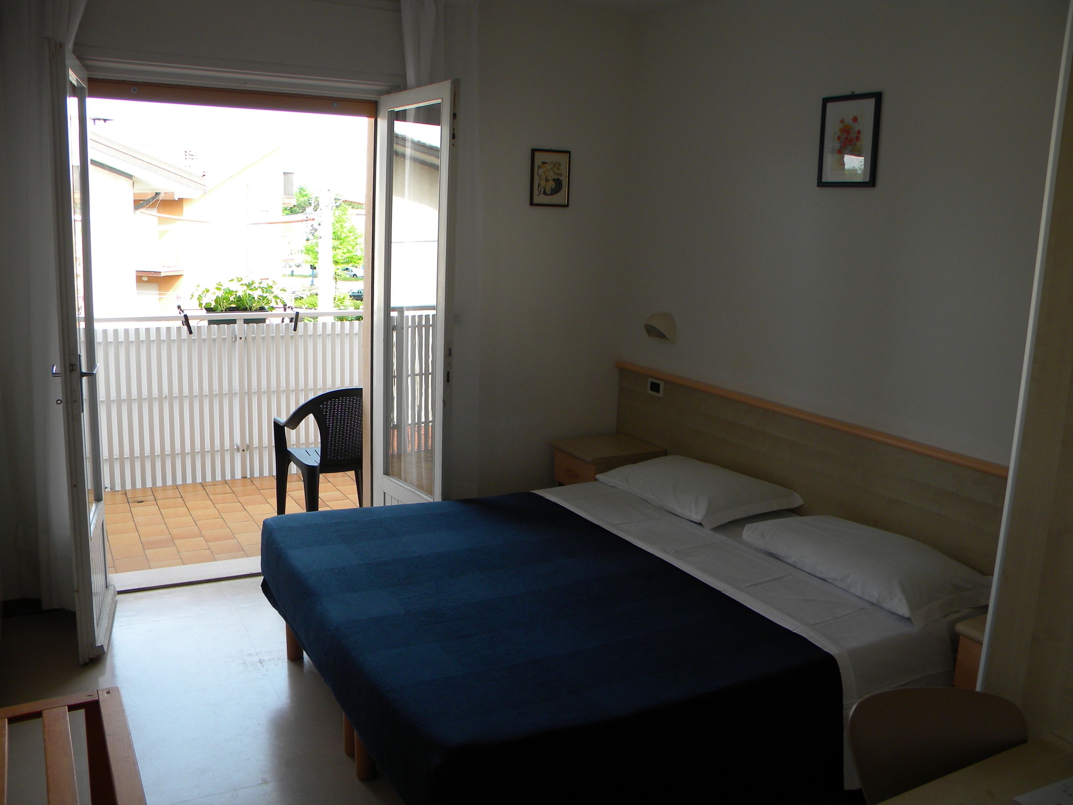 Quarto com cama, uma cadeira e uma pequena mesa. Tem uma varanda com grade branca e uma cadeira lá fora.