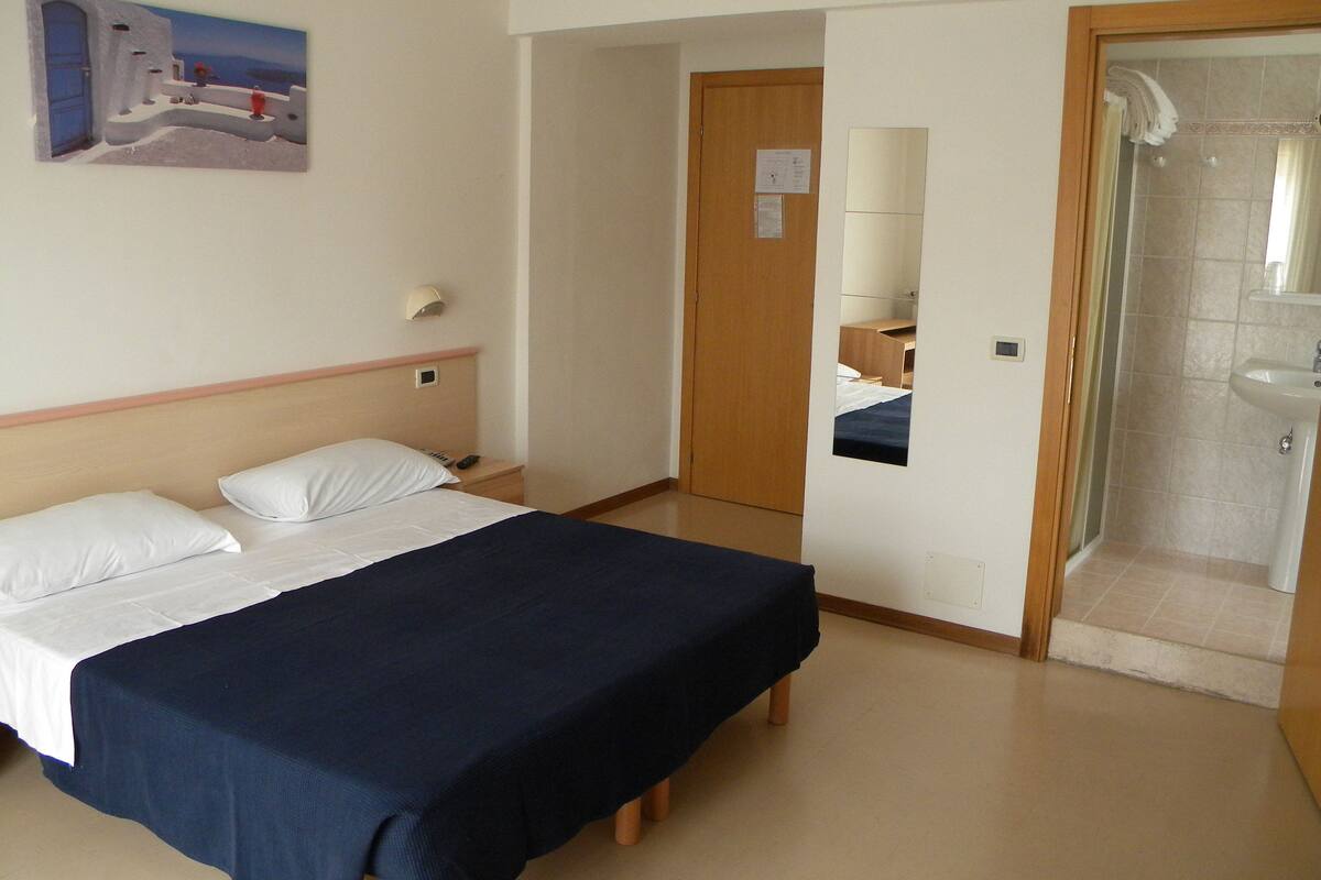Habitación de hotel con cama, mesita de noche, espejo y zona de ducha.
