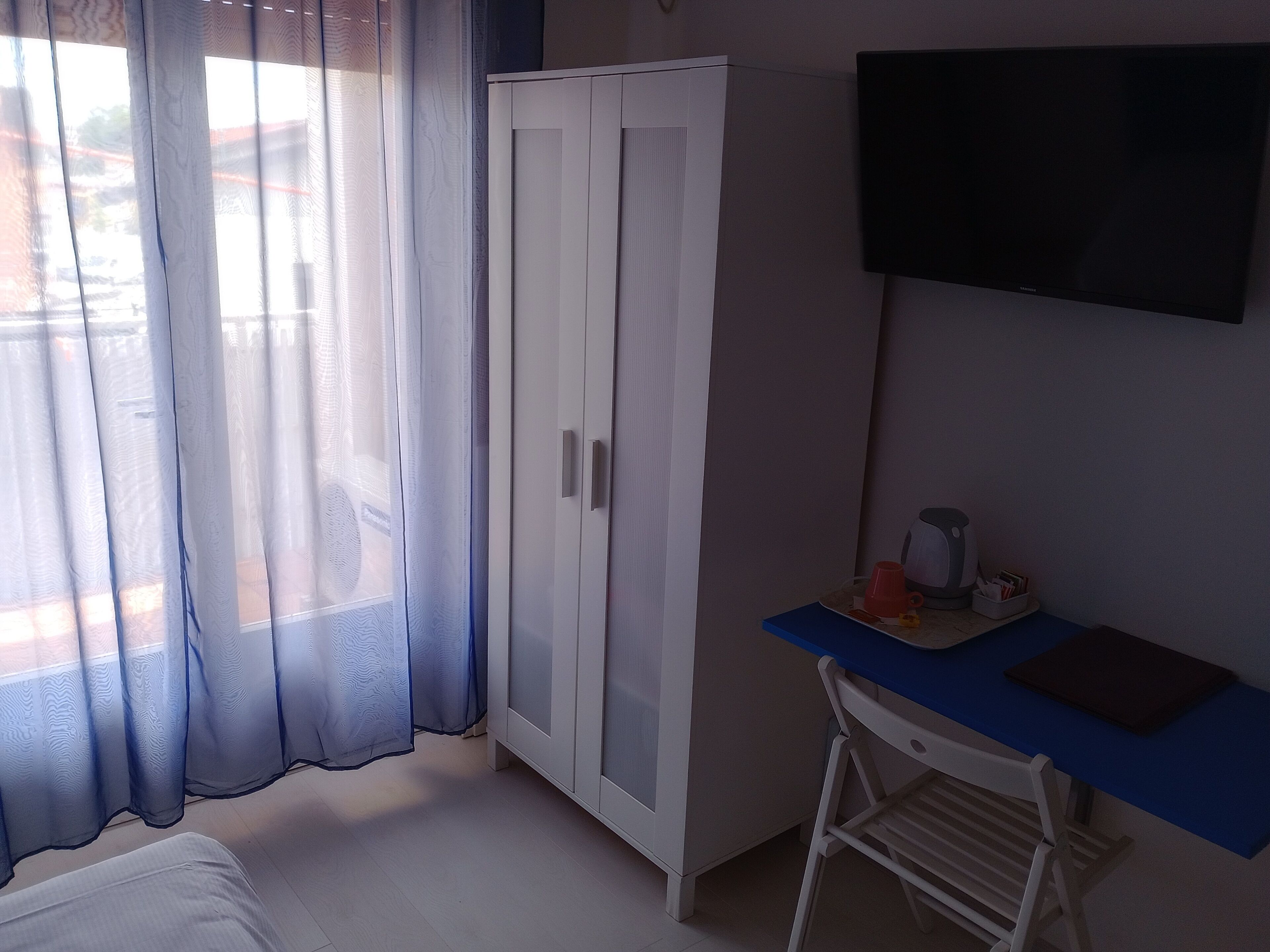 Una habitación con armario, escritorio y silla, televisor y ventana con cortinas translúcidas.