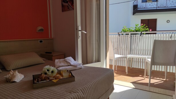 Ein Hotelzimmer mit Bett, einem kleinen Tisch mit Frühstücksangeboten, einem Stuhl und einem Balkon mit weißer Brüstung.