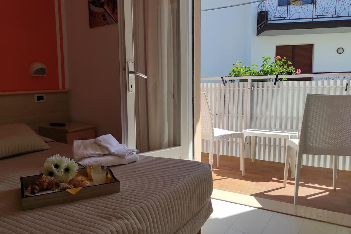 Habitación de hotel con cama, una pequeña mesa con productos para el desayuno, una silla y un balcón con valla blanca.