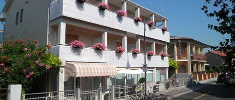 A two-story building with balconies adorned with pink flowers, a visible entrance with a striped awning, and bicycles parked outside.