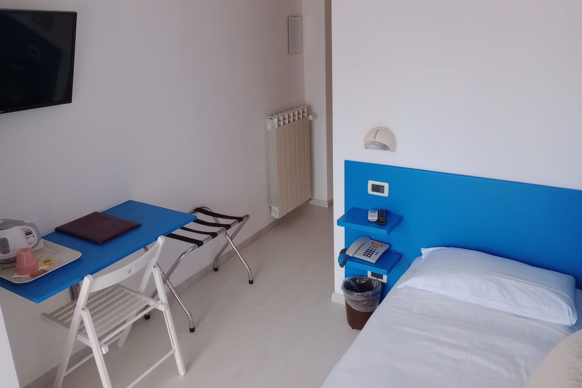 Habitación de hotel con cama, escritorio con silla, teléfono, cesto de basura y radiador.