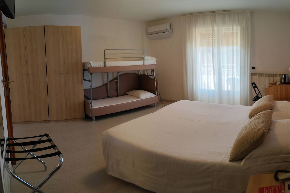 Habitación de hotel compacta con literas, cama individual, armario, aire acondicionado, ventana con cortinas, mesita de noche con lámpara y teléfono.