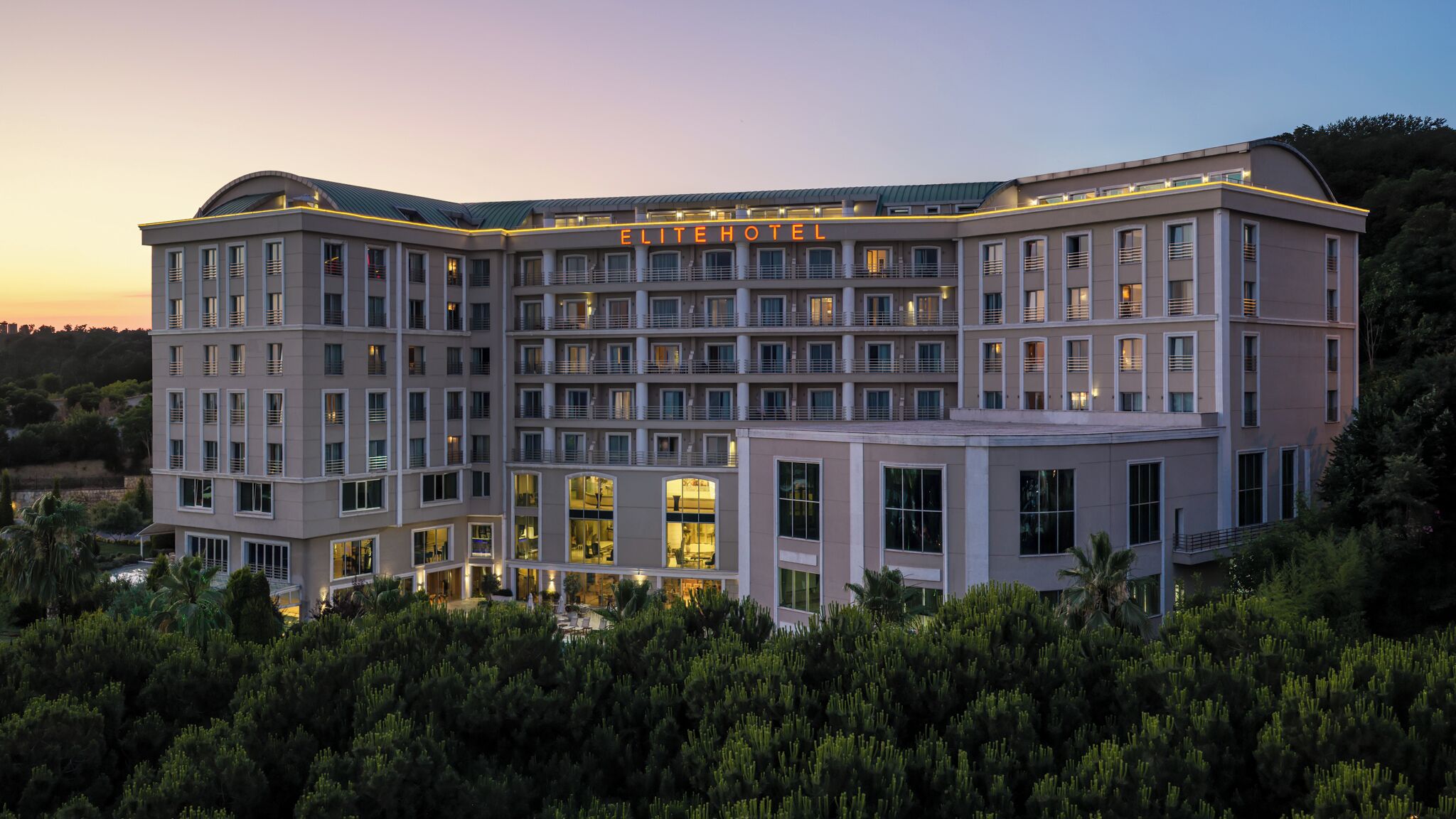 Ein mehrstöckiges Hotel mit einer auffälligen 'ELITE HOTEL'-Beschilderung, großen Fenstern und einer begrünten, angelegten Grünfläche.