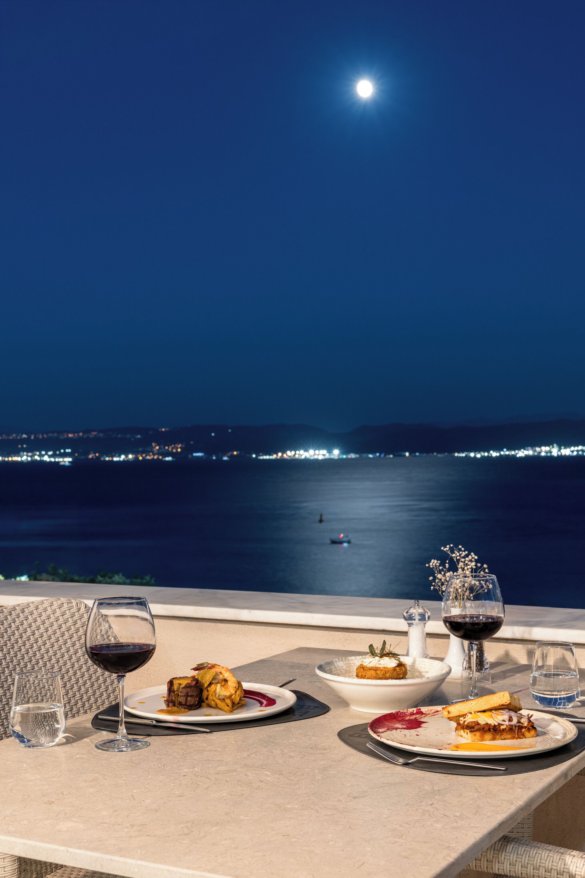 Ein Esstisch, gedeckt für ein Abendessen, mit Blick auf das Meer und eine Stadtlandschaft im Hintergrund.