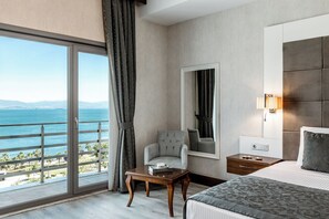 Une chambre d’hôtel avec un lit, une chaise, une table, une table de chevet, un miroir et une vue sur la mer.