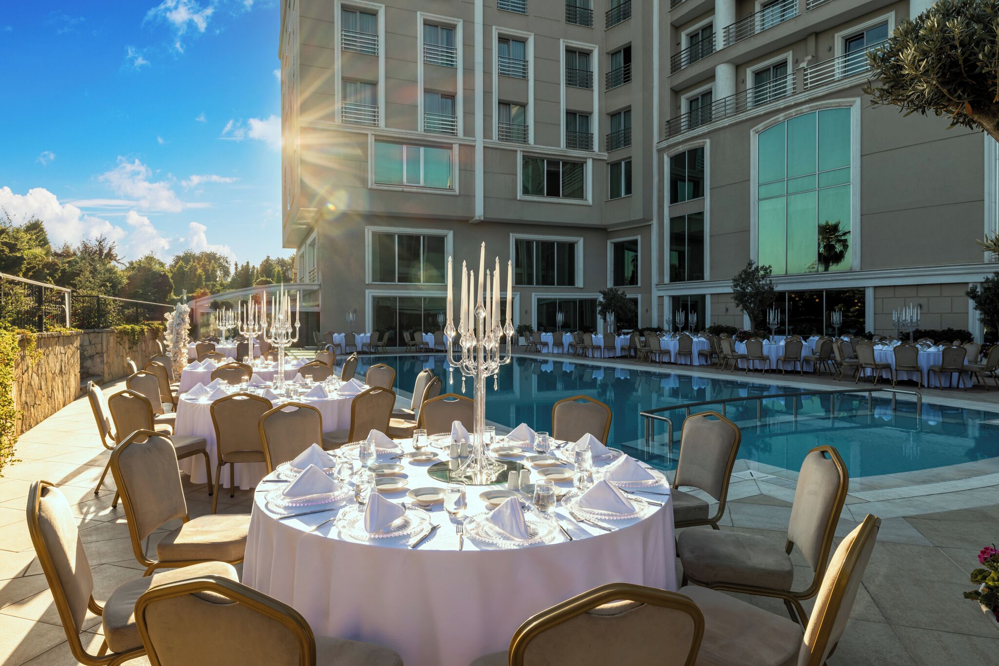 Un área de comedor junto a la piscina con mesas redondas preparadas para una comida, rodeada por una piscina y un edificio con varias ventanas.
