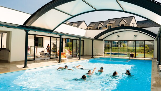 Piscina interior com teto de vidro, vista clara para o exterior e área da piscina com espreguiçadeiras e espreguiçolo.