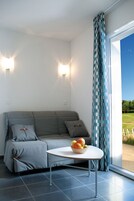 A modern room with a grey sofa, a round table with a fruit bowl, and a view of a green landscape through a large window with patterned curtains.