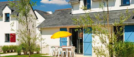 A white house with a grey roof, a patio with a yellow umbrella, and a small garden.