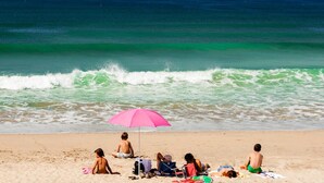 Uma cena de praia com pessoas relaxando sob um guarda-sol rosa, algumas em toalhas e outras sentadas na areia.