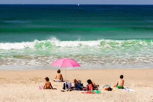A beach scene with people relaxing under a pink umbrella, some on towels and others sitting on the sand.