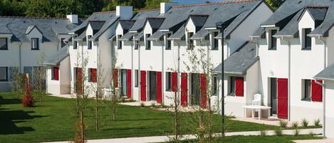 A row of white buildings with red shutters and grey roofs, arranged in a linear fashion with a green lawn in between.