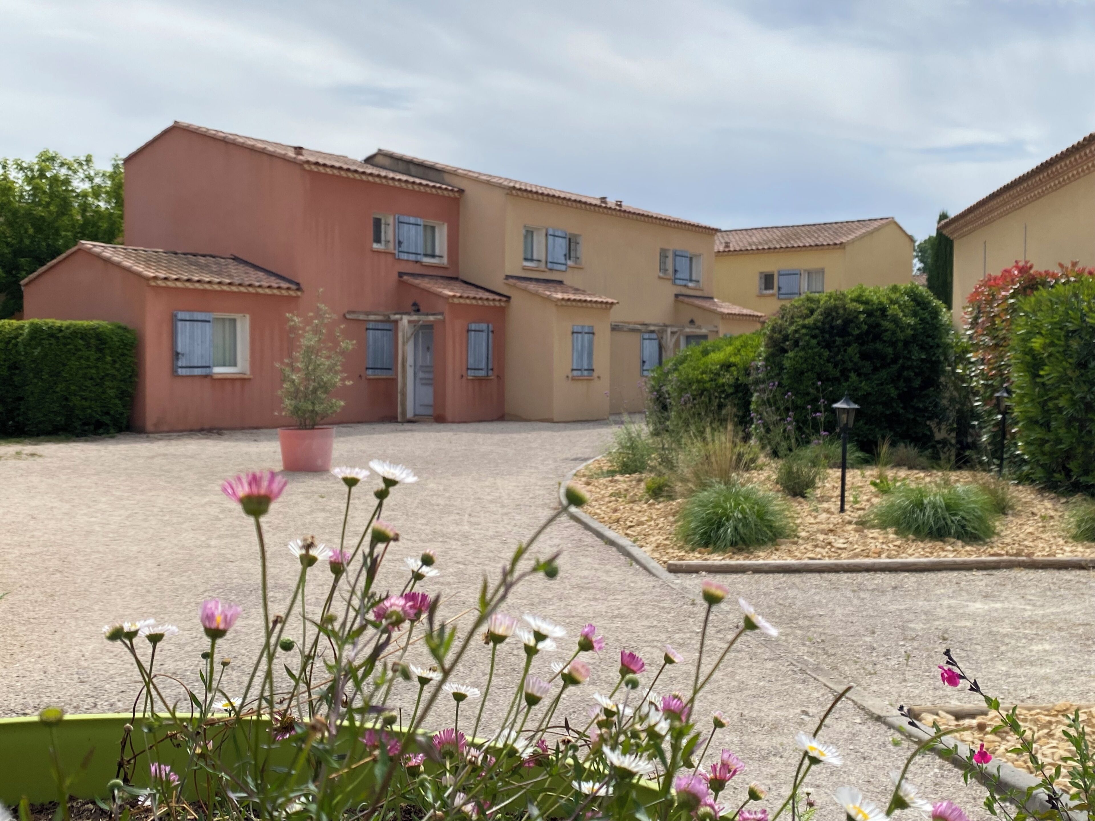 A residential building with multiple balconies, a gravel courtyard, and a variety of flowering plants.