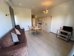 A living room with a brown sofa, a dining table with chairs, a kitchen area, and a TV on a stand. - Résidence Lagrange Vacances L'Oustau de Sorgue (L'Isle-sur-la-Sorgue)