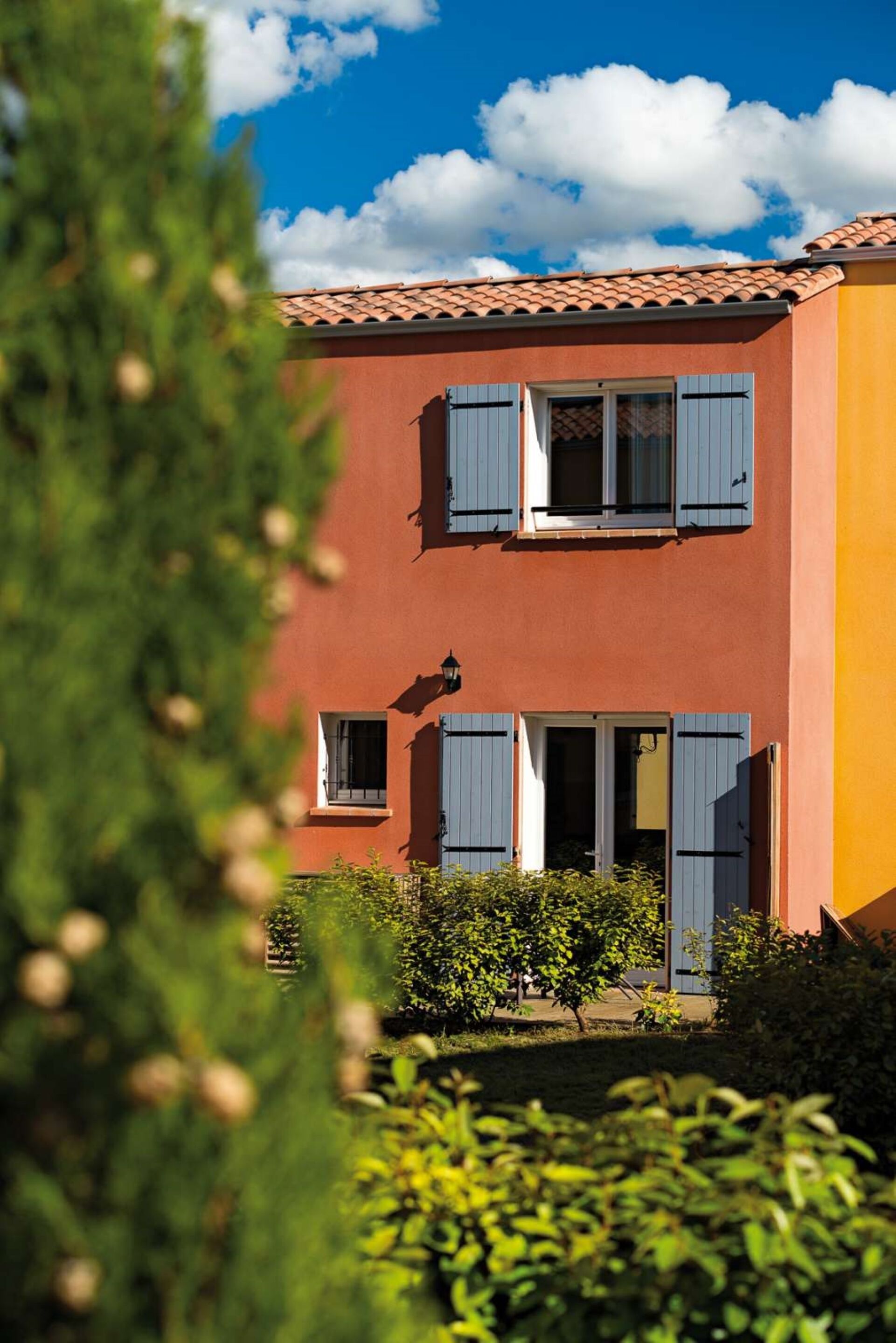 A two-story building with a red facade and blue shutters, surrounded by greenery.