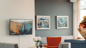A modern lounge area with grey sofas, a wooden coffee table, and a red chair. There are framed artworks on the wall and a large TV screen. - Geo Hotel Grindavik (Grindavik)
