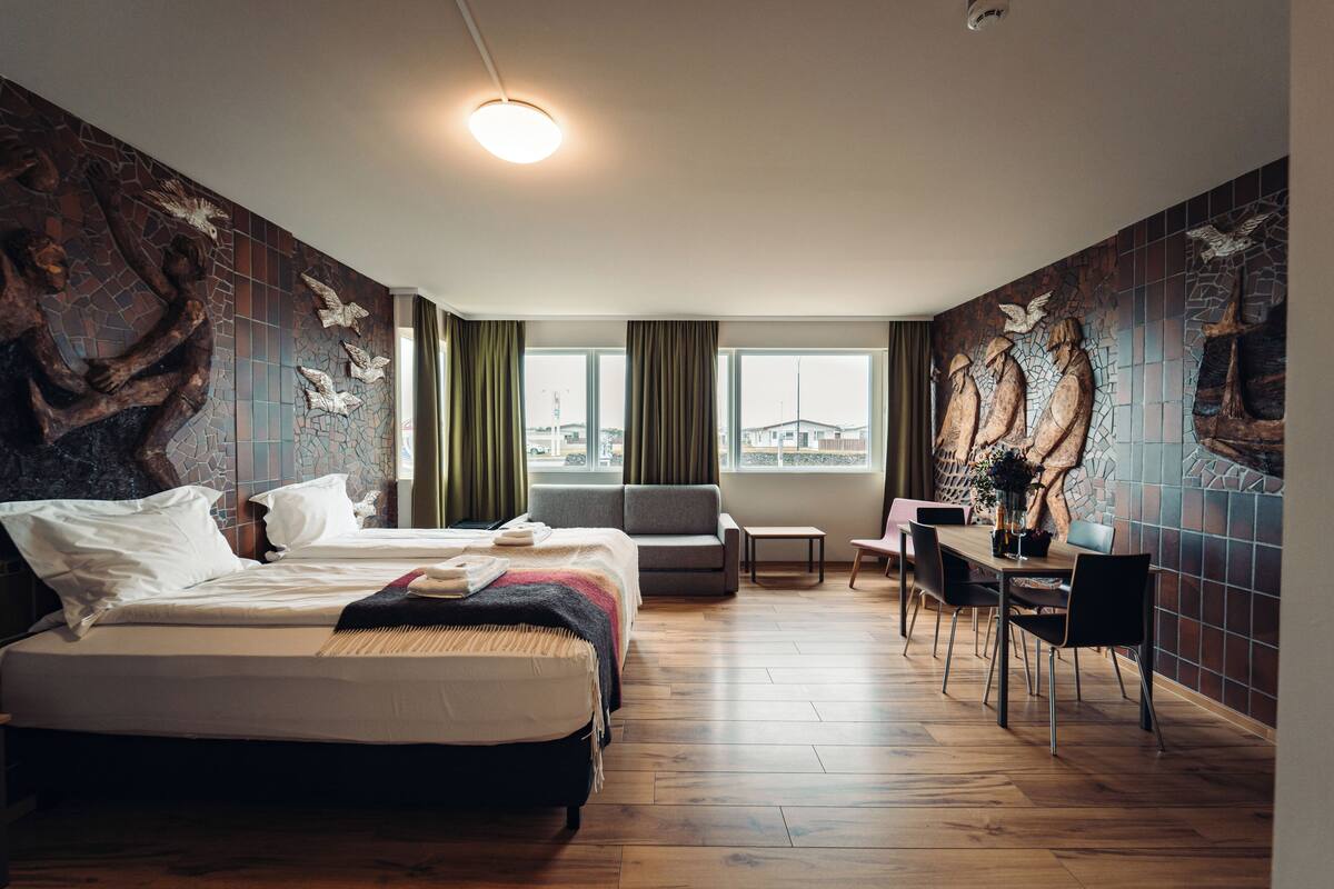 A hotel room with a large bed, a sofa, a desk, and a view of the city through large windows.