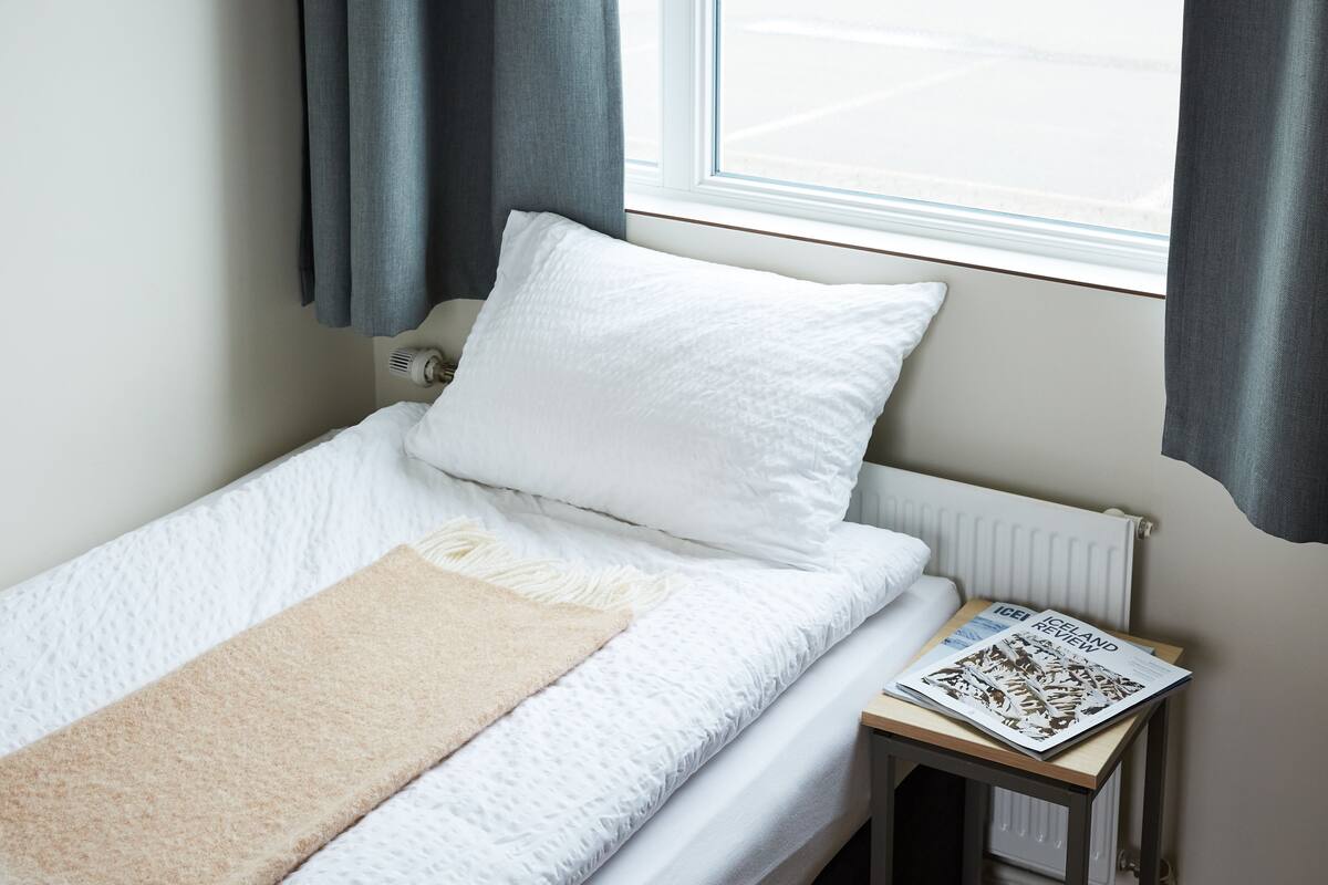 A small, neatly made bed with white bedding and a beige throw blanket, a white pillow, and a magazine on a small table beside the bed.