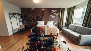 A modern hotel room with a large bed, a grey sofa, a wooden floor, a wall mural, and a window with curtains. - Geo Hotel Grindavik (Grindavik)