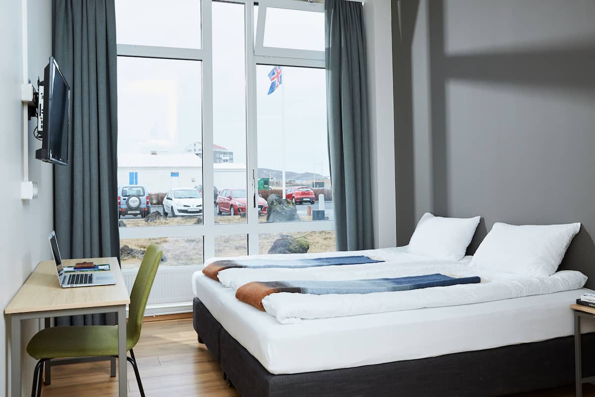 A hotel room with a bed, a desk with a laptop, a chair, and a view of parked cars and a flag outside.