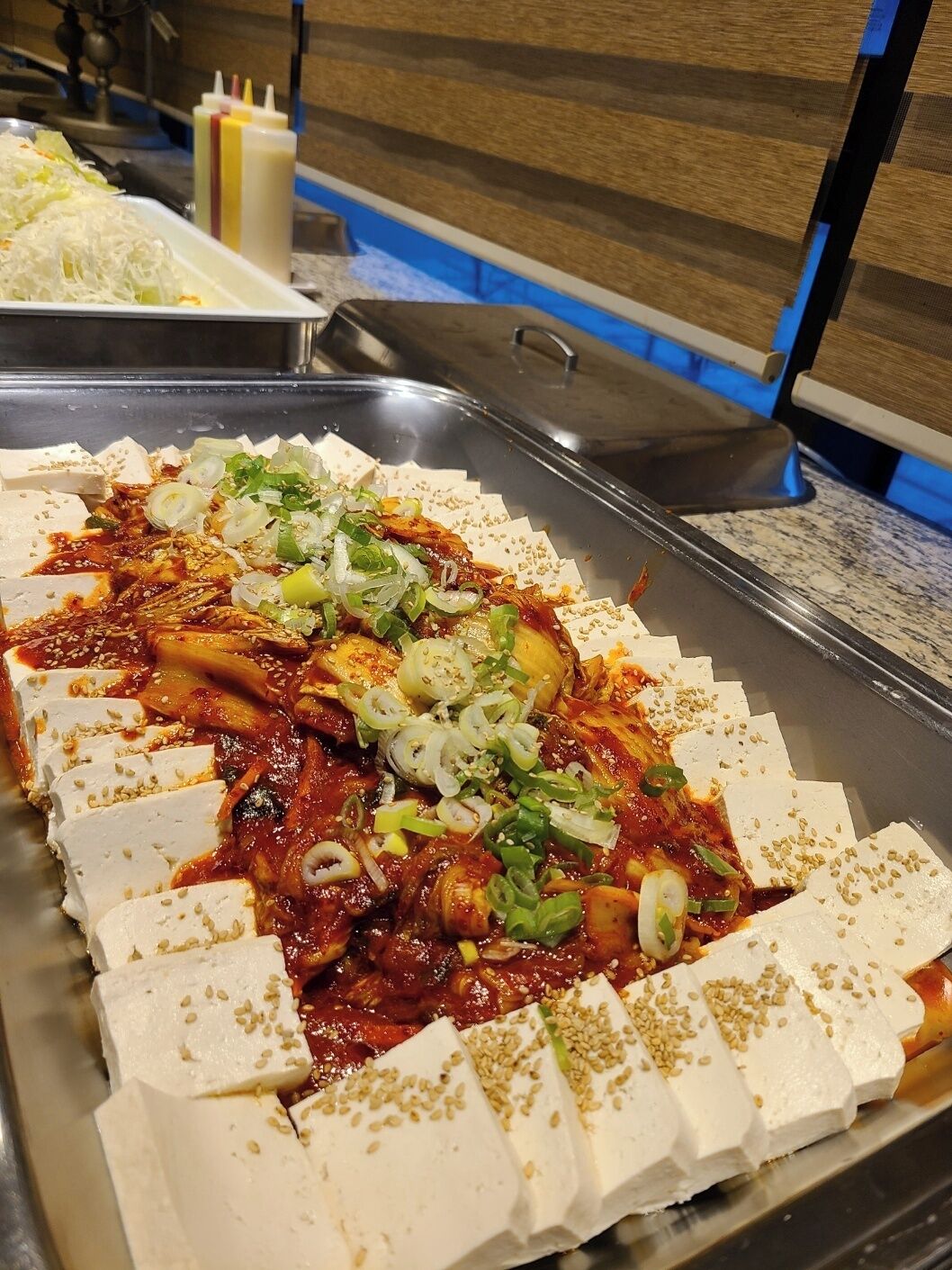 A buffet with various dishes, including a section with tofu and a red sauce, garnished with green onions and sesame seeds.