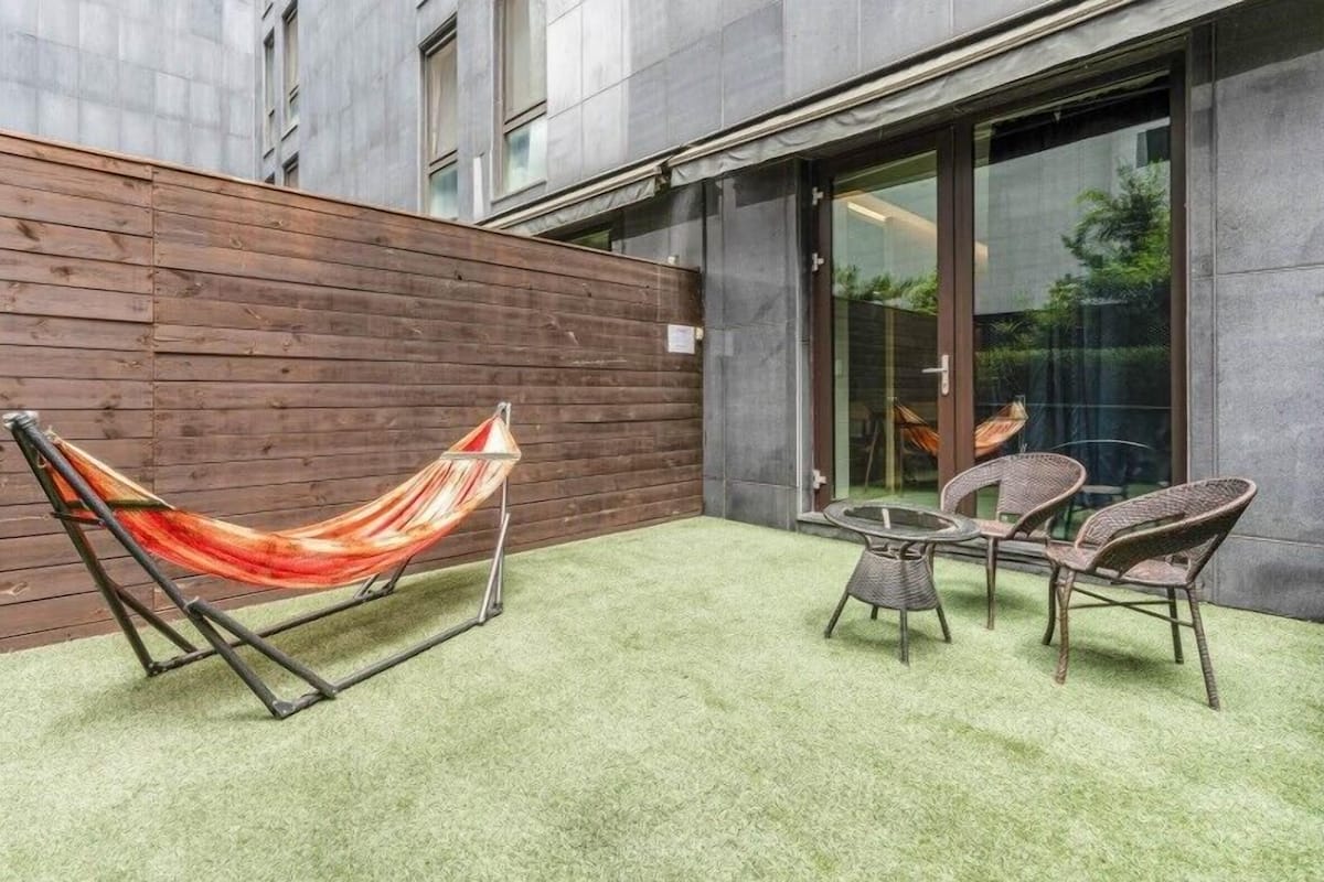 Een hangmat en twee rieten stoelen op een groen gazon met een houten wand en een glazen deur.