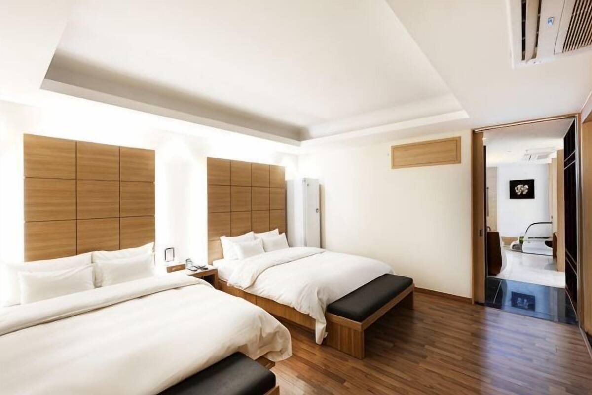 Een hotelkamer met twee bedden, houten hoofdeinden, een bankje, een kledingkast en een zichtbare badkamer door een geopende deur.