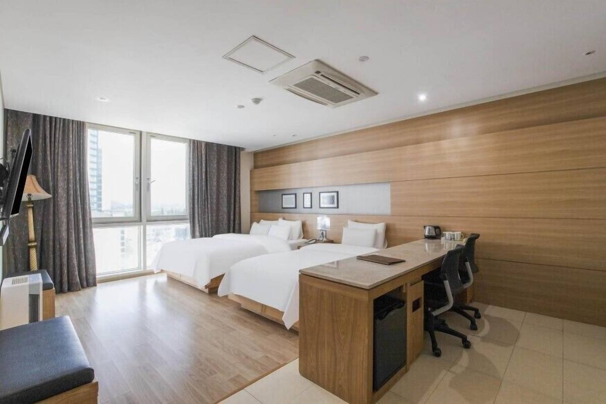 Een hotelkamer met twee bedden, een bureau, een stoel, een lamp en uitzicht op de stad door grote ramen.