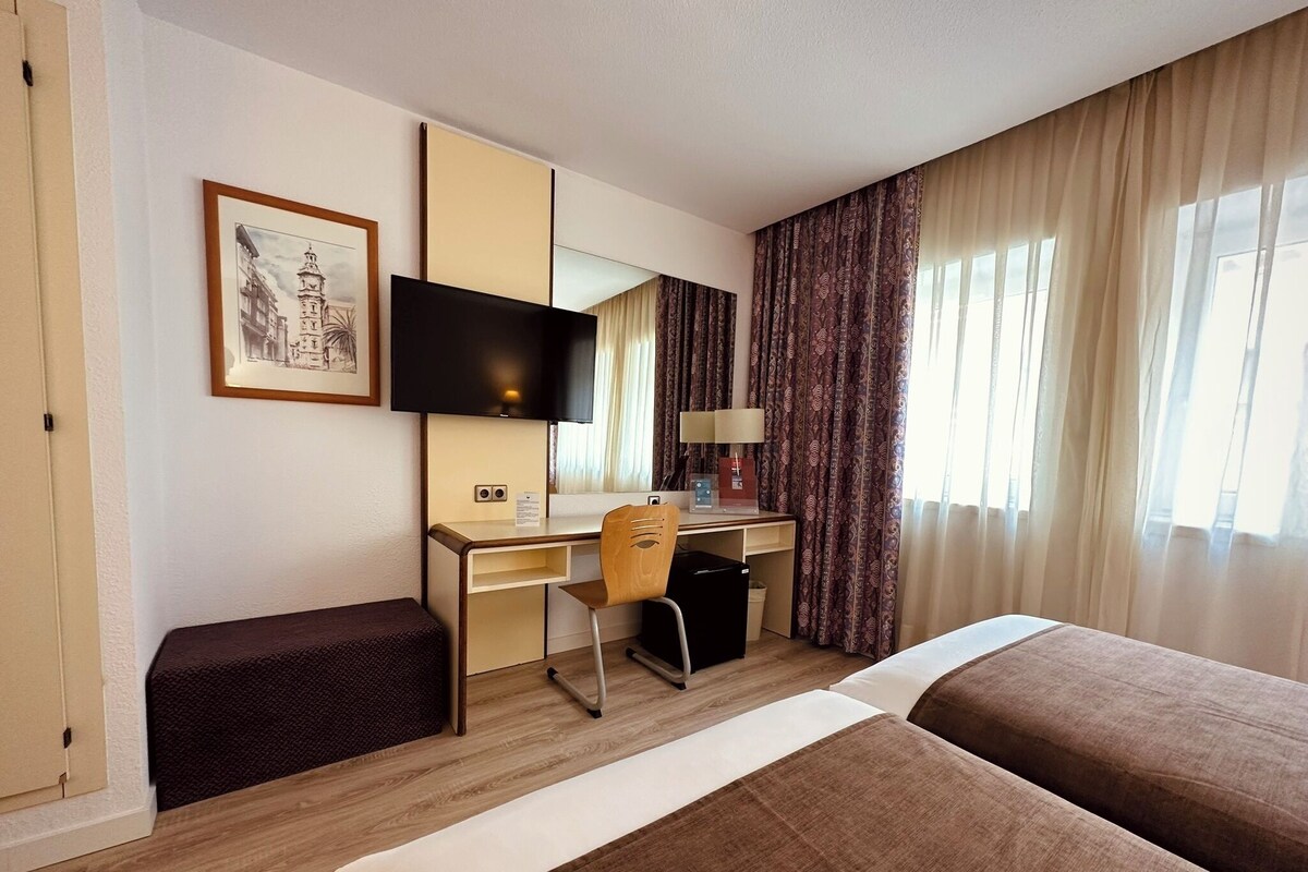 Une chambre d’hôtel comprenant un lit, un bureau, une chaise, une télévision, une fenêtre avec des rideaux et un tableau encadré au mur.