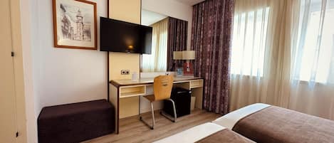 Habitación de hotel con cama, escritorio, silla, televisor, ventana con cortinas y un cuadro en la pared.