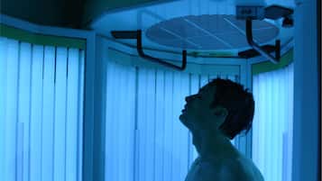 Una persona de pie en una habitación con un ventanal grande, persianas y un ventilador de techo, además de una lámpara.