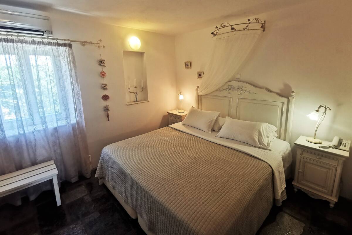 Quarto bem arrumado com cama, mesas de cabeceira, abajur, janela com cortinas e decoração nas paredes.