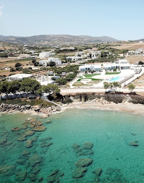 An aerial view of a coastal resort with clear turquoise waters, a swimming pool, and white buildings. - Ambelas Mare (Paros)
