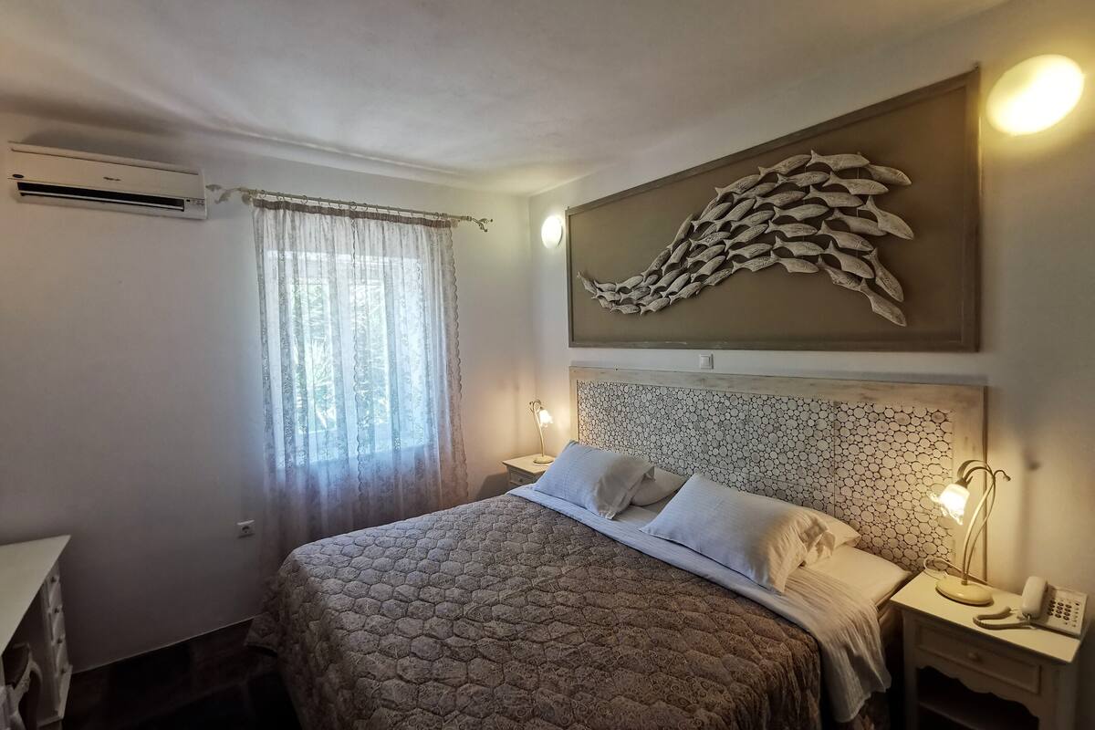 Quarto com cama, mesa de cabeceira, abajur e ar-condicionado fixado na parede.