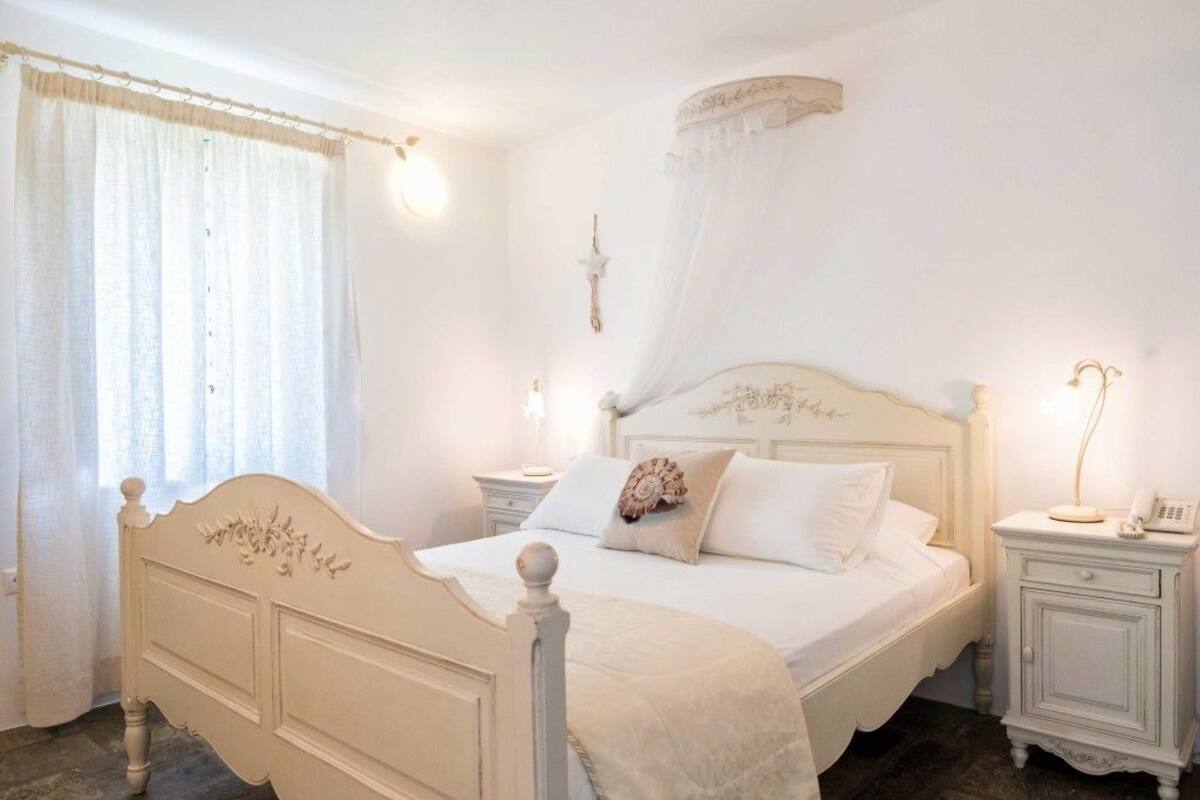Quarto com cama branca, mesas de cabeceira e janela com cortinas.