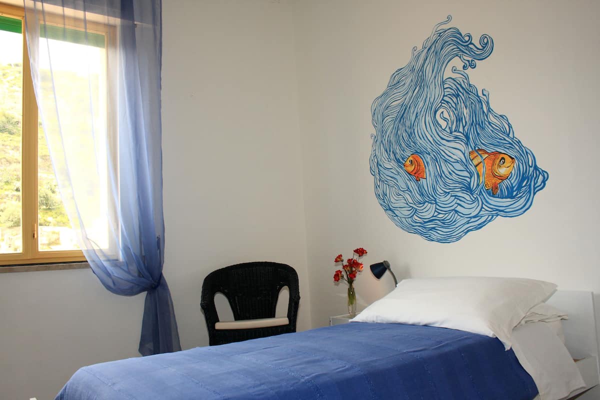 A bedroom with a bed, a chair, a wall art piece, and a window with curtains.