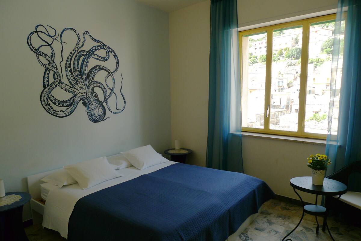 A bedroom with a bed, a wall mural, a window with curtains, a small table with a plant, and a patterned floor.
