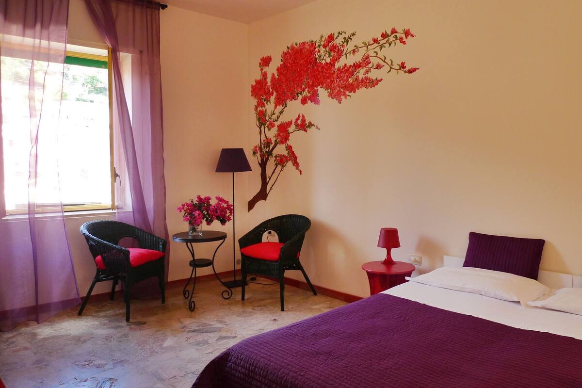 A hotel room with a bed, two chairs, a small table with a vase of flowers, a lamp, and a wall mural.
