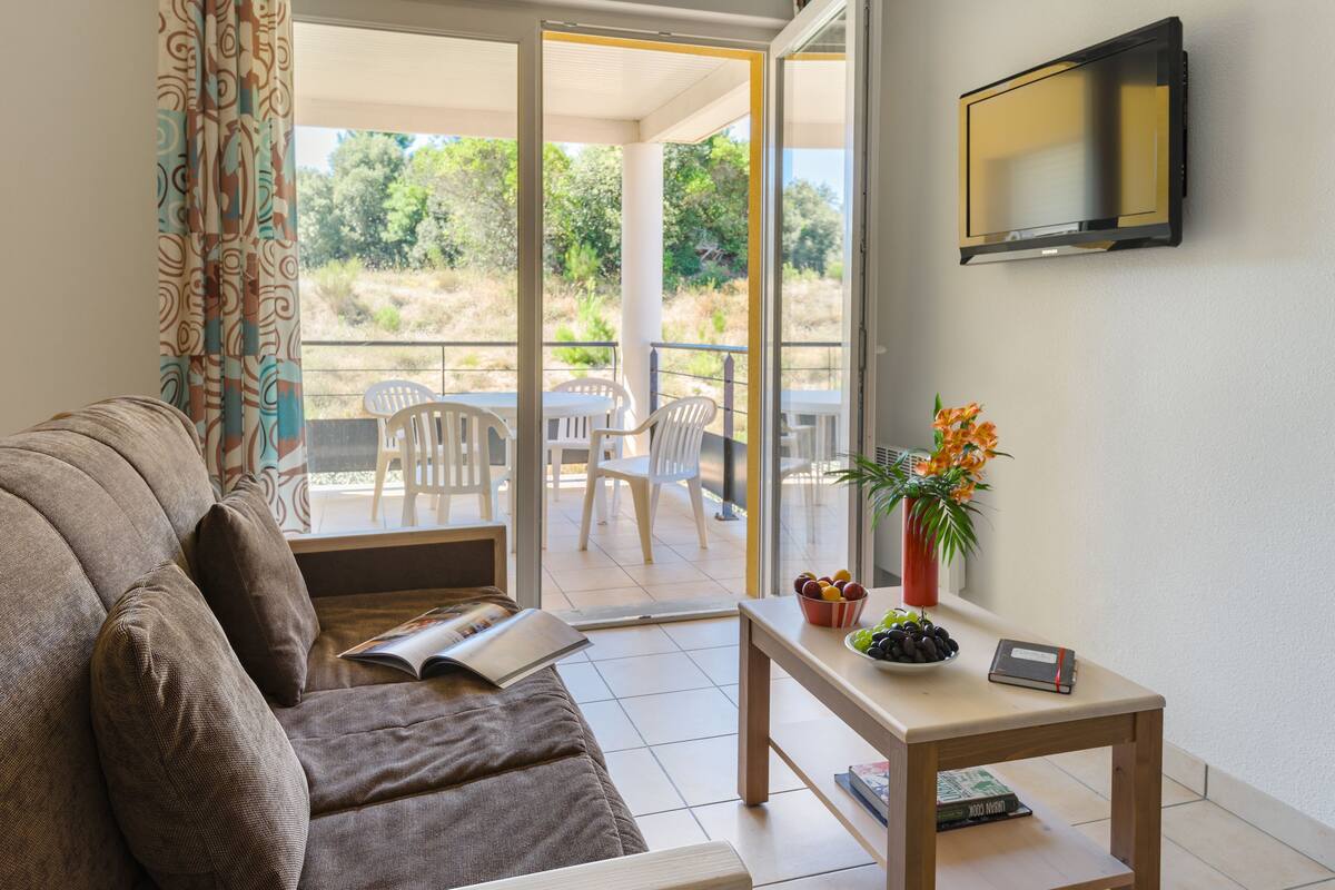 A living room with a sofa, a table with a vase and books, a TV on the wall, and a balcony with chairs and a view of greenery.