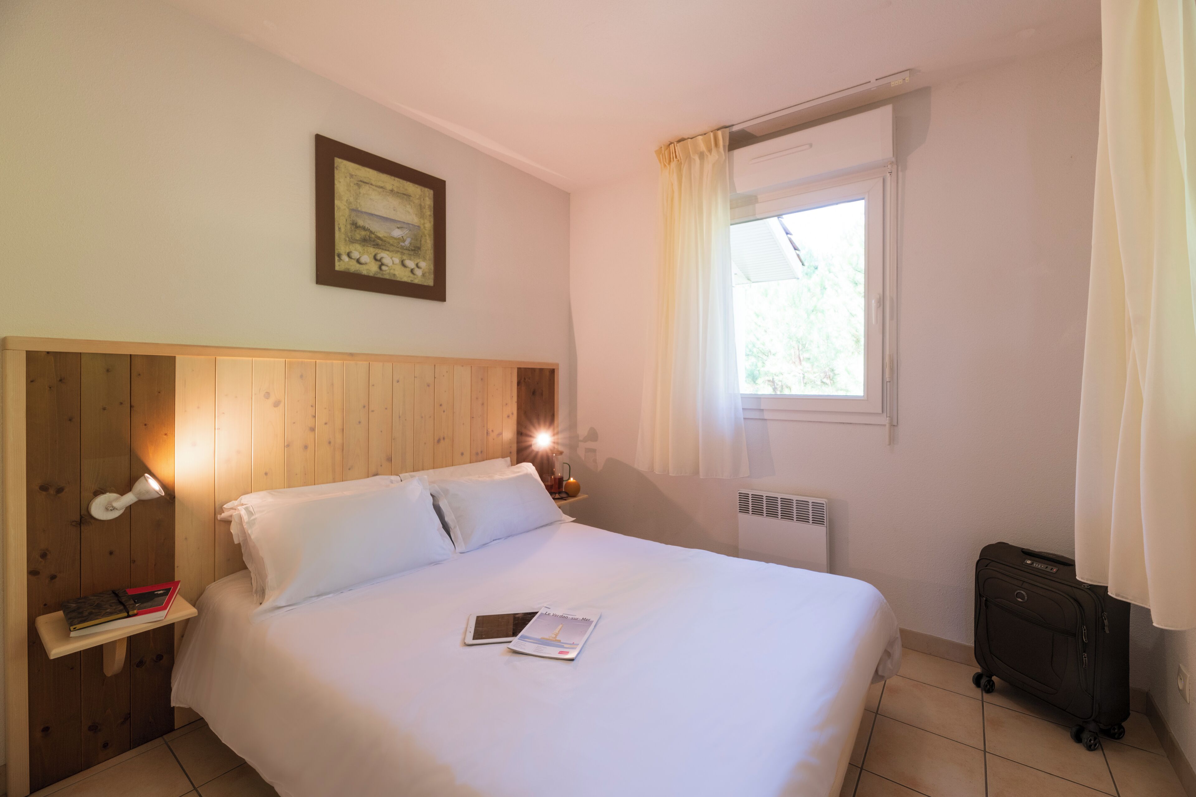 Une chambre d’hôtel avec une tête de lit en bois, une table de chevet avec une lampe, une fenêtre avec des rideaux et une valise.