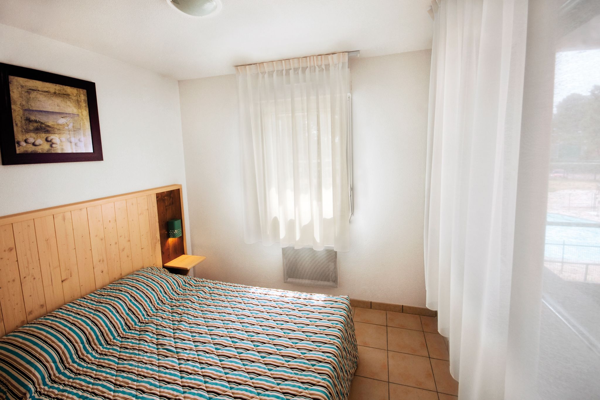 A bedroom with a wooden headboard, a striped bedspread, a small bedside table with a lamp, a window with sheer curtains, and a framed picture on the wall.