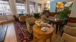 A cozy lounge area with a round wooden table set for tea, comfortable seating, and a large window with blinds.
