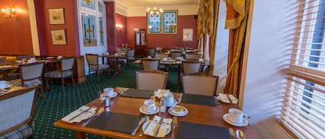 Een restaurantgedeelte van een hotel met meerdere tafels gedekt voor het ontbijt, inclusief stoelen, servies en een groot raam met jaloezieën.
