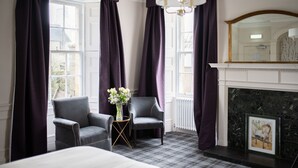 A hotel room with a bed, two armchairs, a small table with a vase, a fireplace, and large windows with purple curtains.