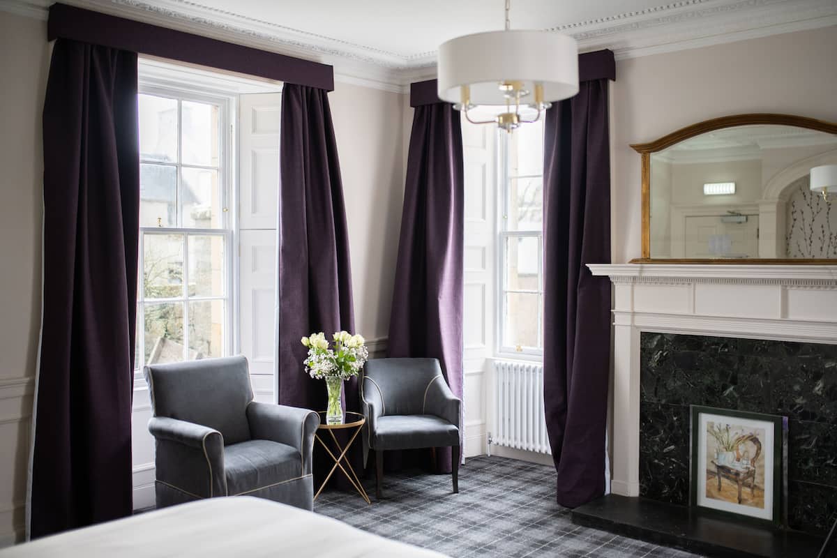 A hotel room with a bed, two armchairs, a small table with a vase, a fireplace, and large windows with purple curtains.